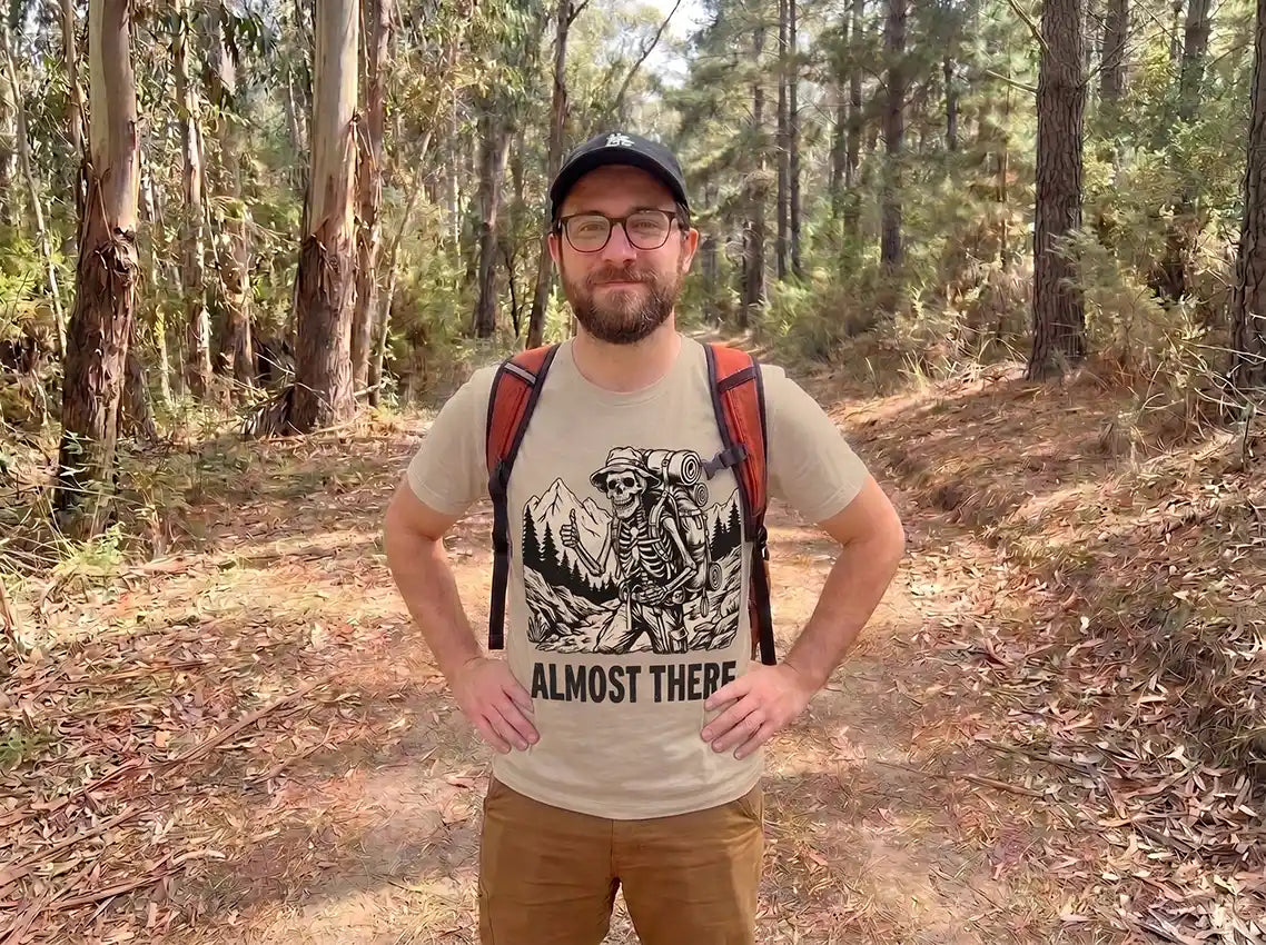 A man wearing a beige graphic t-shirt with a skeleton and mountain design and the text 'ALMOST THERE' stands on a forest path. The t-shirt features a black graphic of a skeleton holding a backpack with red straps, wearing a black cap and glasses.