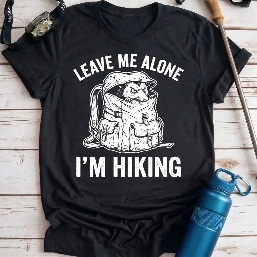 A black short-sleeve graphic T-shirt laid flat on a light wood surface, with white text reading “LEAVE ME ALONE I’M HIKING” and a white line drawing of a dog sitting inside a hiking backpack. Hiking props including a watch, trekking pole, and blue water bottle are placed around the shirt.