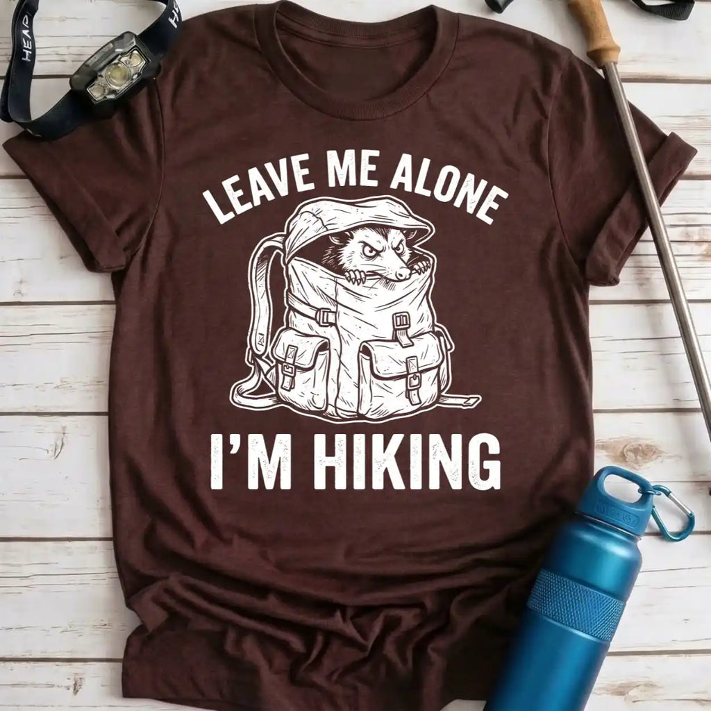 A brown short-sleeve graphic T-shirt laid flat on a light wood surface, with white text reading 'LEAVE ME ALONE I'M HIKING' and a hiking dog in a backpack illustration. A blue water bottle, watch, strap, and trekking pole are placed around it as props.