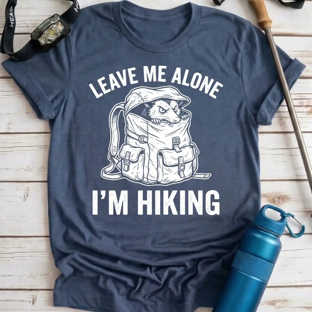 A navy blue graphic t-shirt laid flat on a light wood background, featuring a white line art illustration of a dog peeking out of a hiking backpack with the text 'LEAVE ME ALONE I'M HIKING'. A blue water bottle, watch, and trekking pole are styled around the shirt.