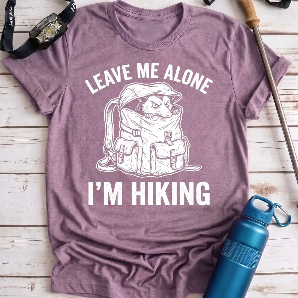 A heathered mauve purple t-shirt with a white graphic of a hiking dog in a backpack and the text 'LEAVE ME ALONE I’M HIKING'. The t-shirt is laid flat on a light wood background with outdoor gear props including a blue water bottle, a watch, and a trekking pole.