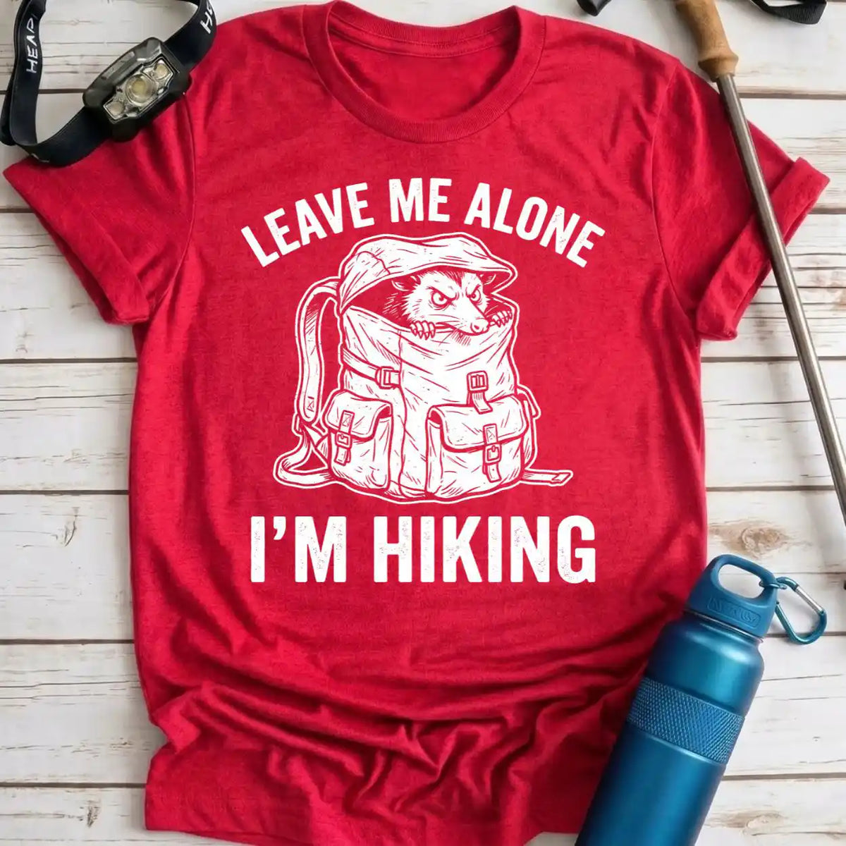 A red short-sleeve graphic T-shirt laid flat on a light wood surface, with white text reading 'LEAVE ME ALONE I'M HIKING' and a white line drawing of a dog wrapped in a backpack. Hiking props including a watch strap, rod, and blue water bottle are placed around the shirt.