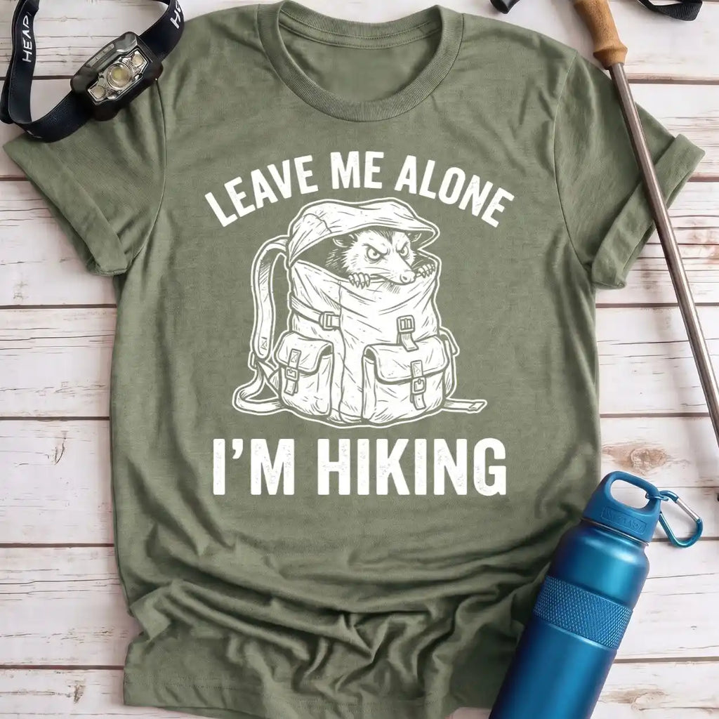 An olive green graphic t-shirt laid flat on a light wood background, featuring a white line art illustration of a dog inside a hiking backpack with the text 'LEAVE ME ALONE I'M HIKING'. Hiking props like a water bottle, watch, and trekking pole surround the shirt.