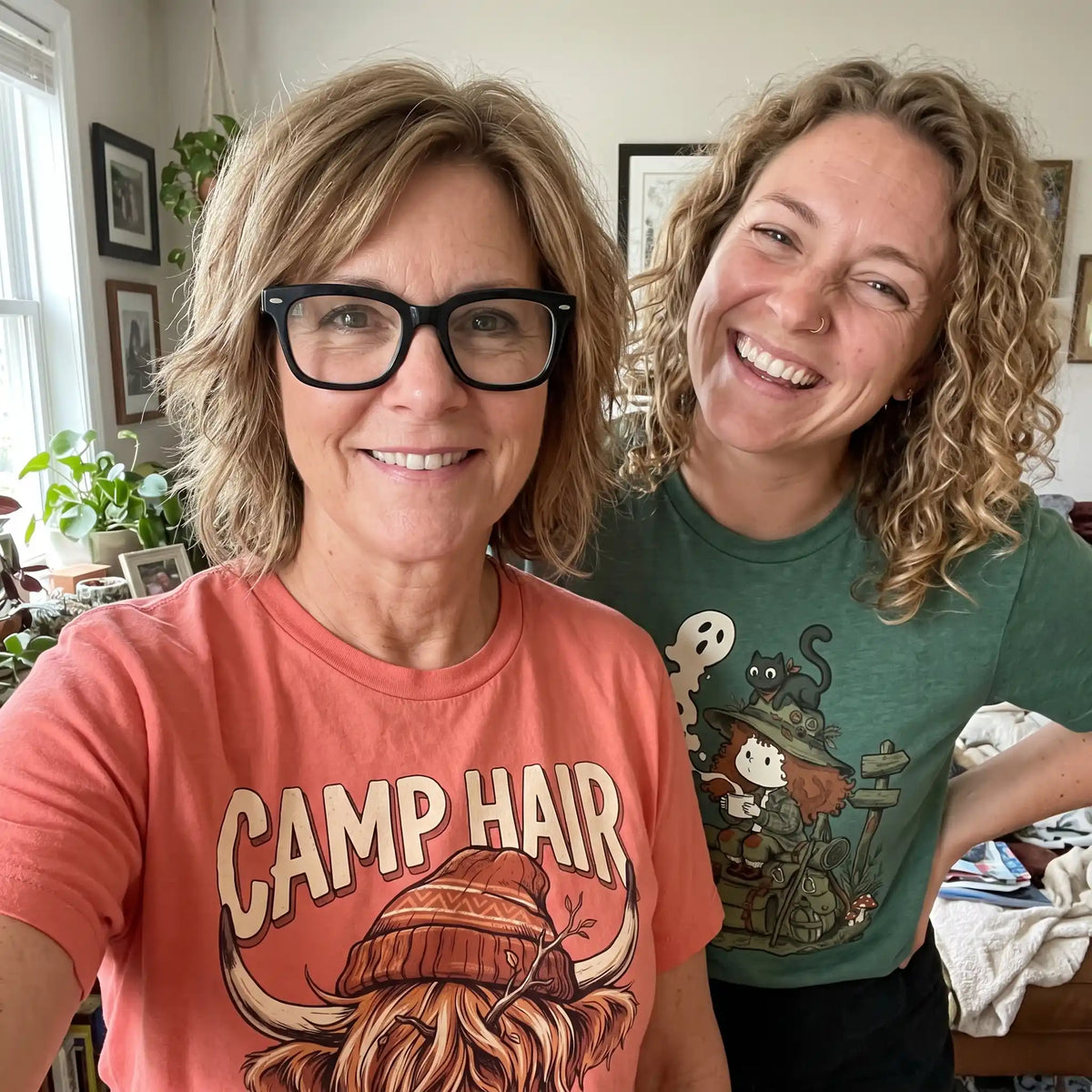 Two women smiling for a selfie in a home setting. The woman on the left wears a coral-pink graphic t-shirt with 'CAMP HAIR' and a long wavy hair illustration, while the woman on the right wears a green graphic t-shirt with a witch and cat design.