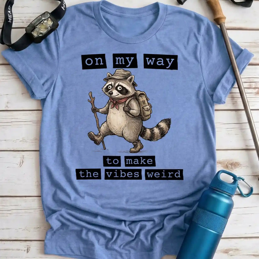 A light blue graphic t-shirt laid flat on a white wood background, featuring a cartoon raccoon wearing a hat and backpack with the text 'on my way to make the vibes weird'. A blue water bottle, watch strap, and trekking pole are placed around it as props.
