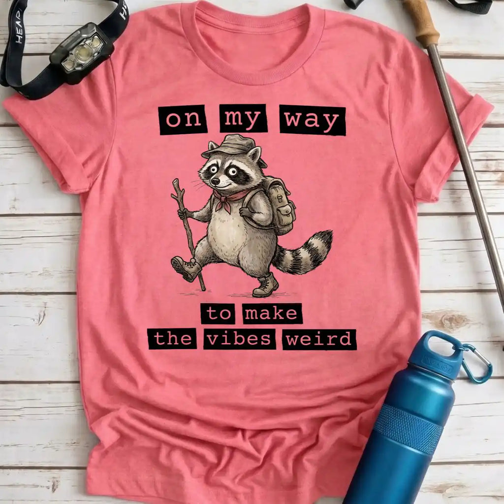 A pink short-sleeve graphic T-shirt laid flat on a whitewashed wood surface, featuring a raccoon hiker illustration with the text 'on my way to make the vibes weird.' A watch, pen, and blue water bottle are placed around the shirt as styling props.