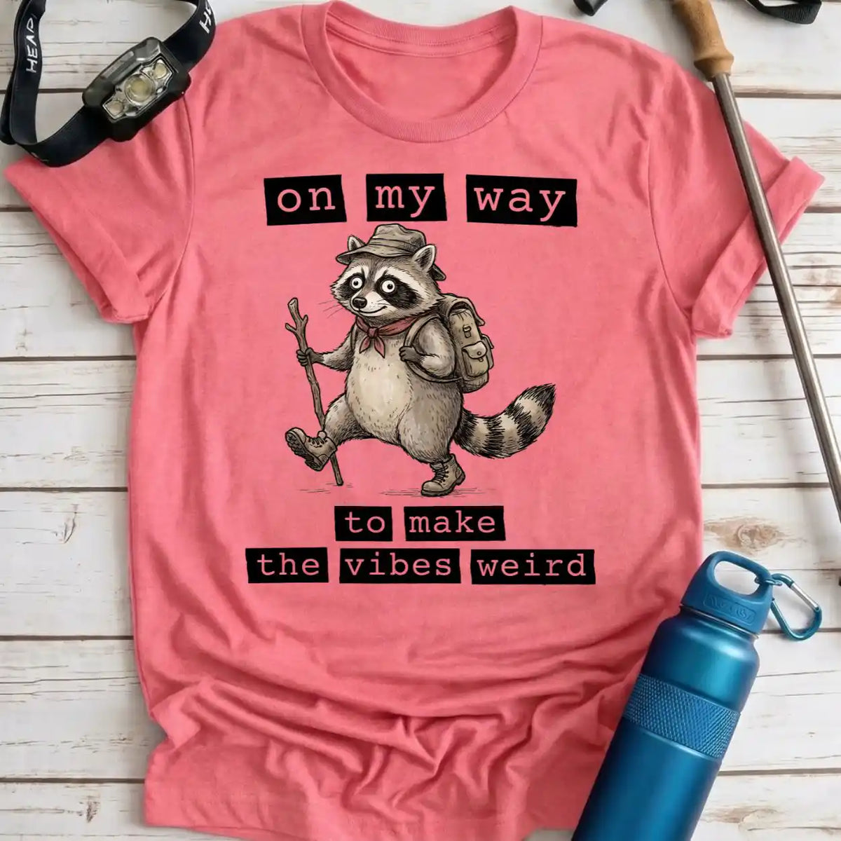 A pink short-sleeve graphic T-shirt laid flat on a whitewashed wood surface, featuring a raccoon hiker illustration with the text 'on my way to make the vibes weird.' A watch, pen, and blue water bottle are placed around the shirt as styling props.