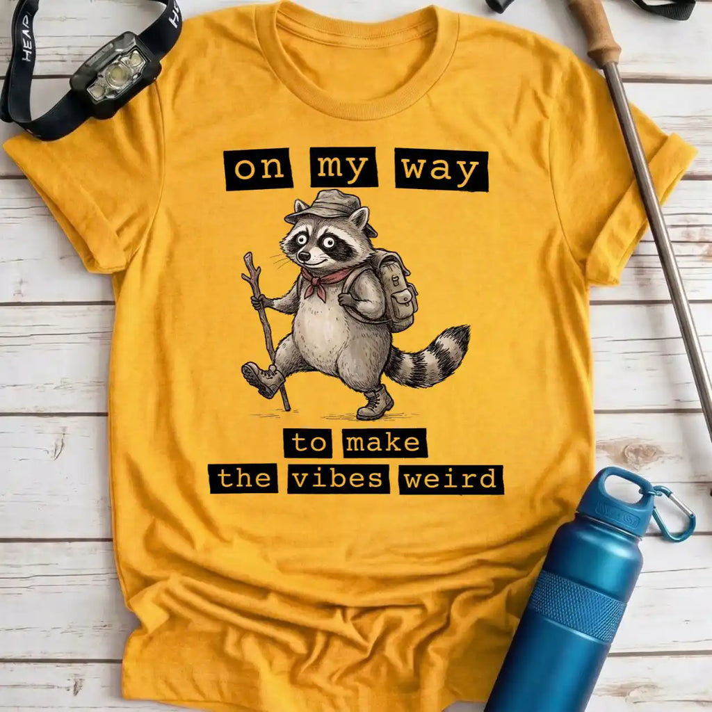 A yellow graphic t-shirt laid flat on a white wood background, featuring a cartoon raccoon wearing a hat and backpack with the text 'on my way to make the vibes weird'. A blue water bottle, watch strap, and trekking pole are placed around it as props.