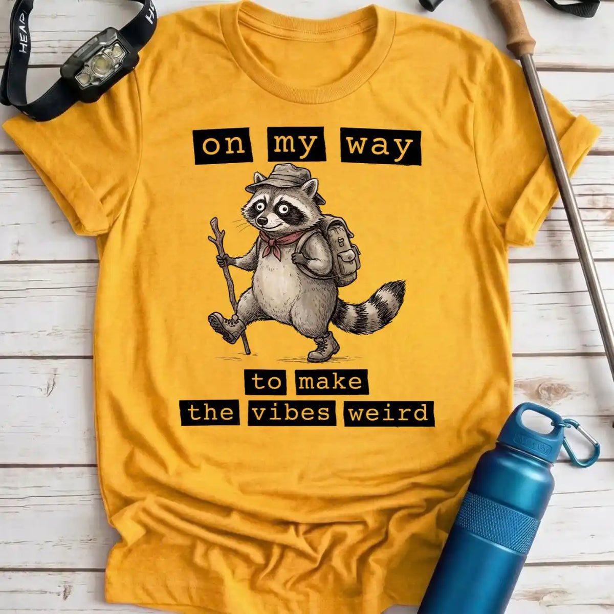 A yellow graphic t-shirt laid flat on a white wood background, featuring a cartoon raccoon wearing a hat and backpack with the text 'on my way to make the vibes weird'. A blue water bottle, watch strap, and trekking pole are placed around it as props.