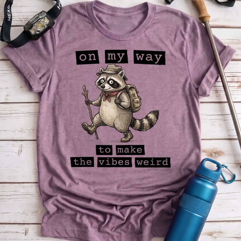 A heathered lavender-purple t-shirt featuring a cartoon raccoon hiker with the text 'on my way to make the vibes weird'. The shirt is laid flat on a white wood plank background, styled with a blue water bottle, a watch, and a trekking pole.