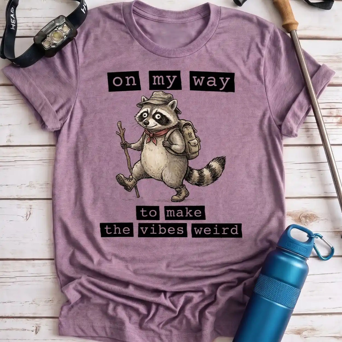 A heathered lavender-purple t-shirt featuring a cartoon raccoon hiker with the text 'on my way to make the vibes weird'. The shirt is laid flat on a white wood plank background, styled with a blue water bottle, a watch, and a trekking pole.