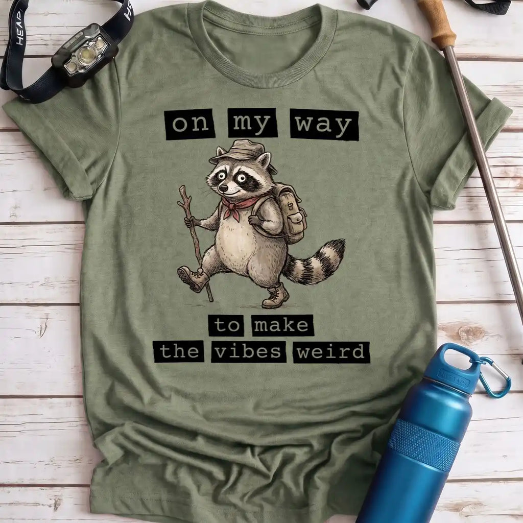 An olive green graphic t-shirt laid flat on a light wood background, featuring a cartoon raccoon hiker with the text 'on my way to make the vibes weird'. A blue water bottle, watch strap, and trekking pole are styled around the shirt.
