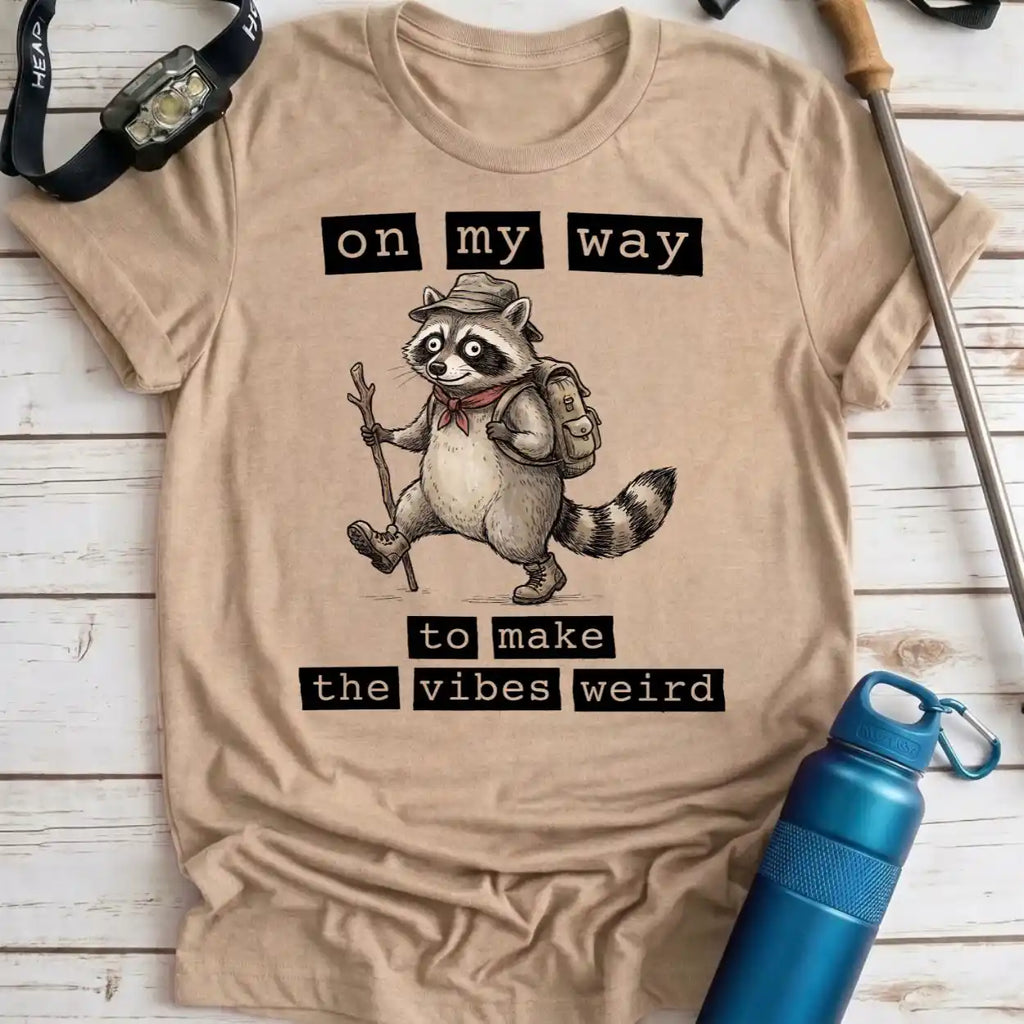 A beige graphic t-shirt laid flat on a white wood plank background, surrounded by outdoor accessories including a blue water bottle, a watch, and a trekking pole. The t-shirt features a cartoon raccoon wearing a hat, backpack, and scarf, with the text 'on my way to make the vibes weird'.