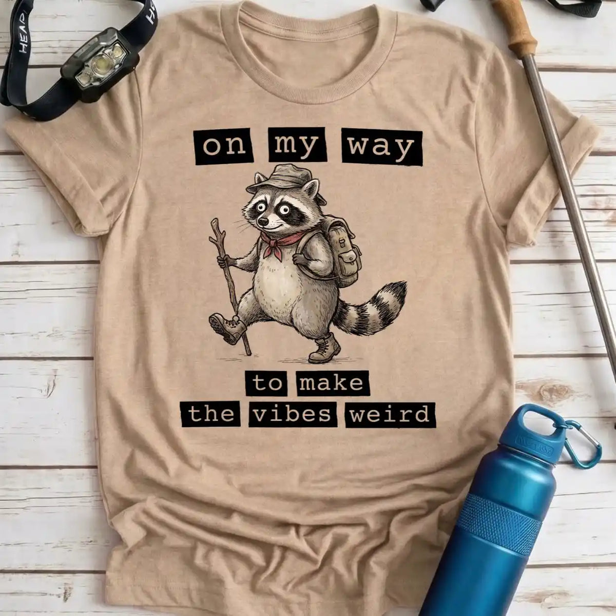 A beige graphic t-shirt laid flat on a white wood plank background, surrounded by outdoor accessories including a blue water bottle, a watch, and a trekking pole. The t-shirt features a cartoon raccoon wearing a hat, backpack, and scarf, with the text 'on my way to make the vibes weird'.
